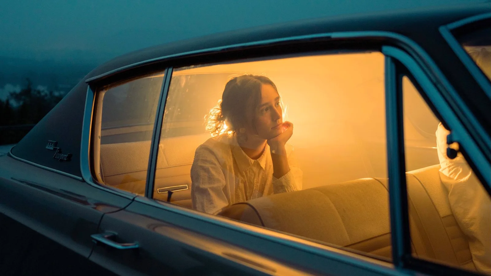 A young woman in a white shirt rests her chin on her hand, glowing orange inside a car seen through its window at dusk.