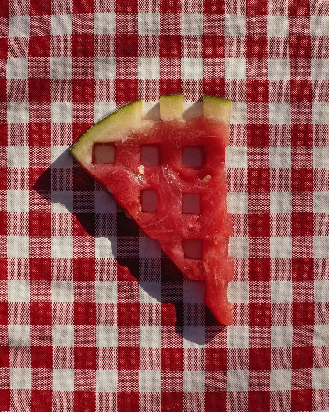 A watermelon wedge with a few square holes, on a red and white gingham cloth, casting a sharp shadow.