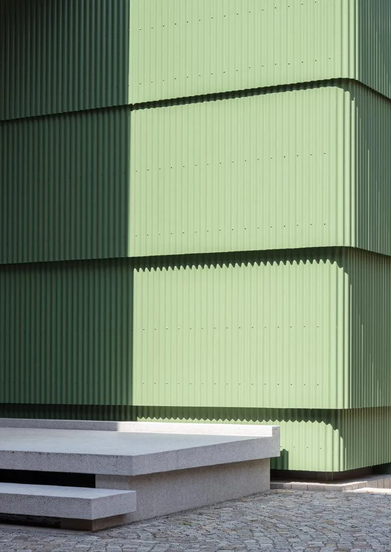 A corrugated sage-green building in stacked tiers, half in shadow and half in sunlight, above a pale concrete platform and cobbled paving.